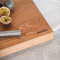Bild in Galerie-Betrachter laden, Hackbrett aus Holz Natur, Auffangöffnung für Teller, Milieubild in Küche, weißer Teller in Auffangöffnung, Messer und Früchte liegen auf Brett, Ecke mit Cookx Aufschrift